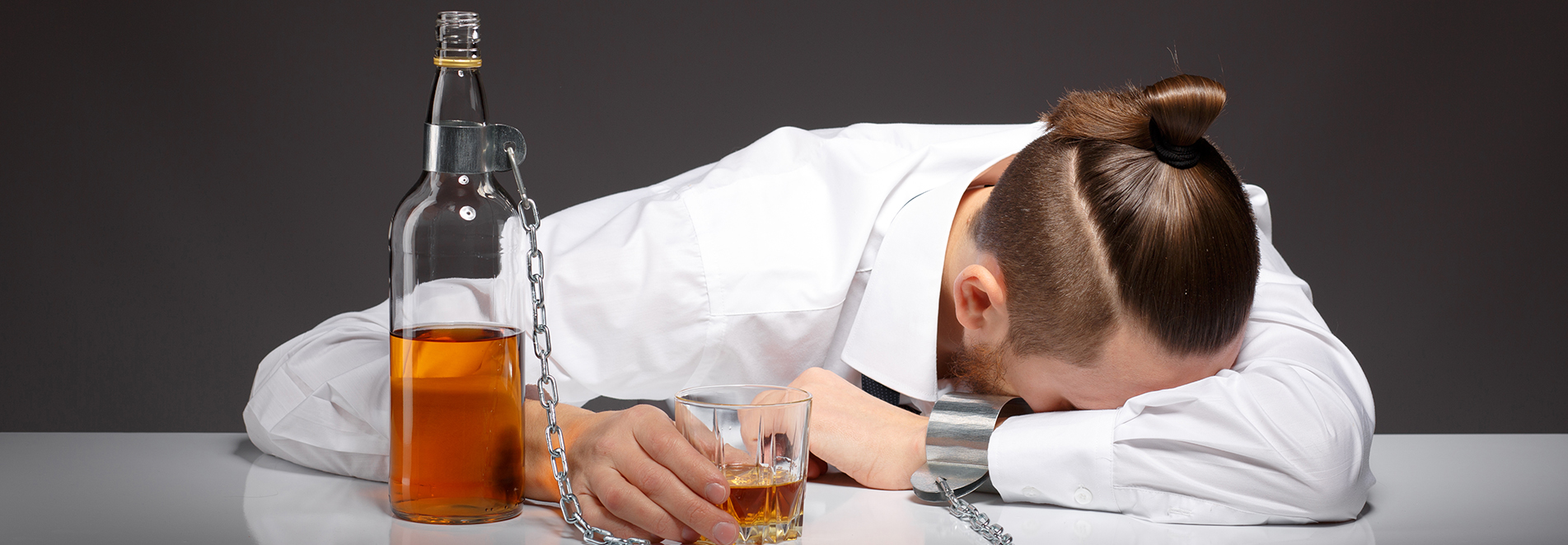 Alcohol Addiction Treatment Surprise, AZ - A mans sits with his head down on a table. His left hand is chained to a bottle of alcohol that sits next to him with a glass of alcohol is in his right hand.