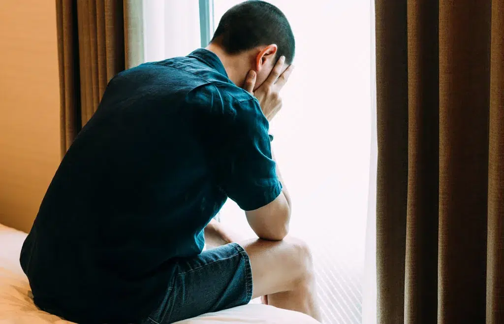 A man feeling broken holds his head in his hands while sitting on the edge of a bed