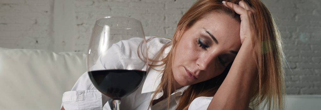 Alcohol Rehab - Close up photo of a woman with a glass of wine in front of her as she holds her left hand on top of her head running her fingers through her hair. She has a depressed look on her face and needs an alcohol rehab to help with her alcoholism.