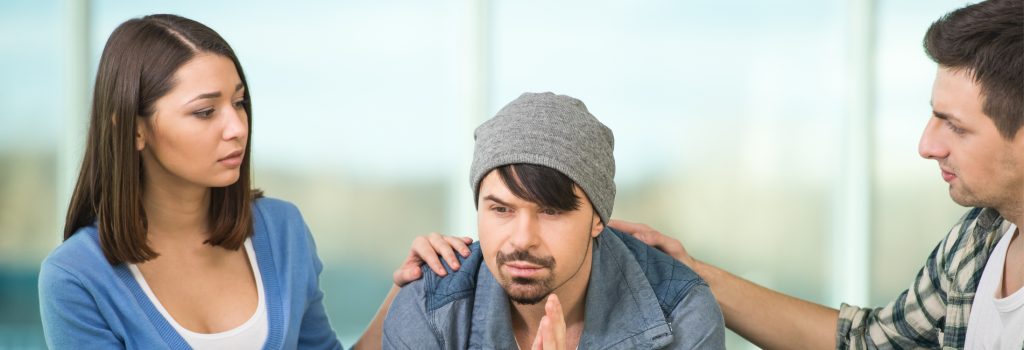  Inpatient Alcohol Rehab - A man sits between an woman and another man who have their hands on his shoulders encouraging him to talk about his alcoholism during group therapy.